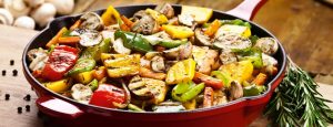 Eine Pfanne mit gebratenem Gemüse steht auf einem Holzbrett.