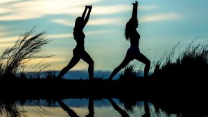Die schwarzen Silhouetten von zwei Frauen, die Dehnübungen vor einem See machen.