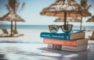 Drei gestapelte Bücher und eine Sonnenbrille liegen am Strand auf einem Tisch, im Hintergrund Sonnenschirme aus Bambus