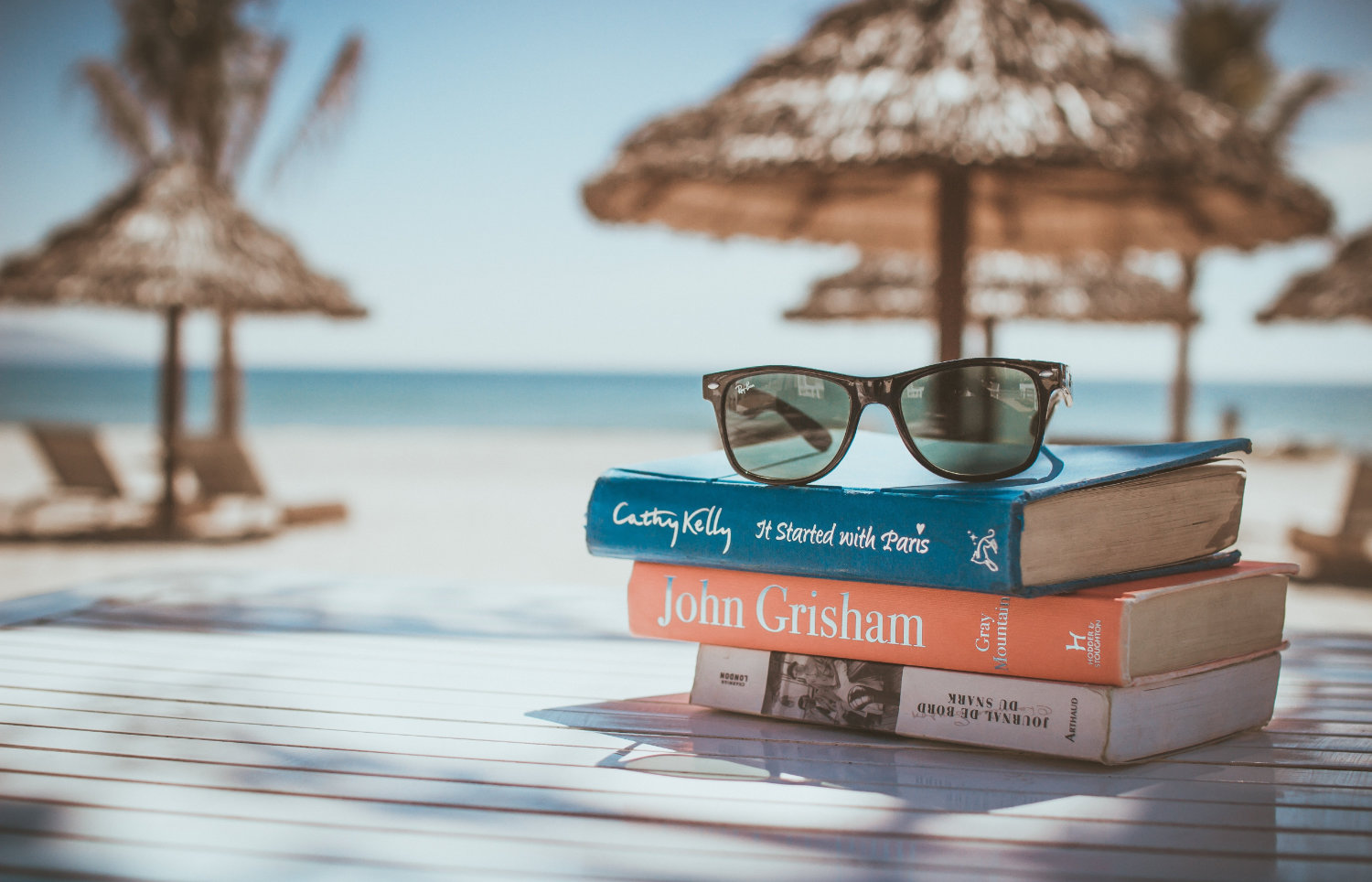 Bücher und Sonnenbrille liegen am Strand auf einem Tisch, im Hintergrund Sonnenschirme aus Bambus