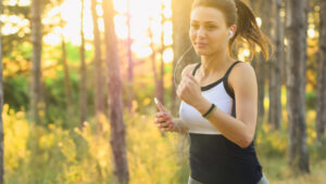 Eine Junge Frau mit Handy in der Hand und Kopfhörern in den Ohren joggt durch einen Wald.