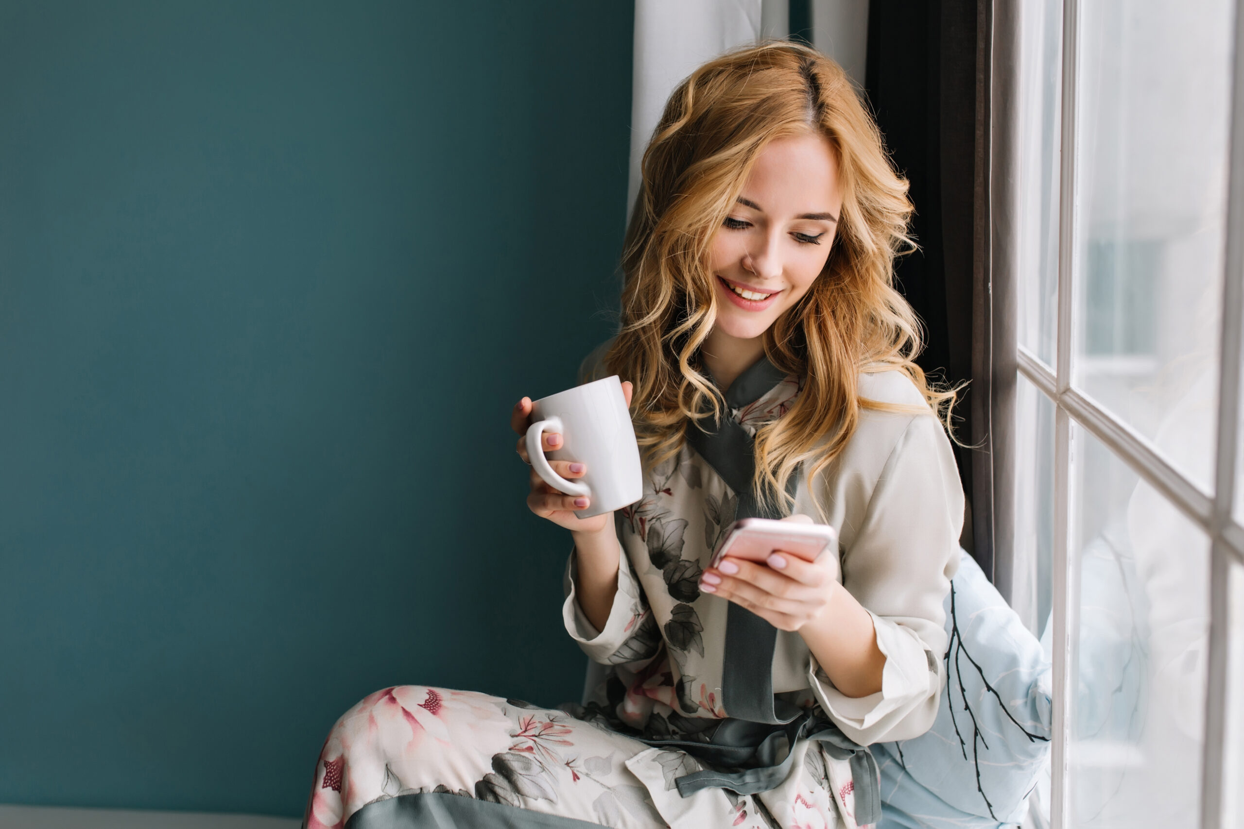 Hübsches blondes Mädchen sitzt auf der Fensterbank mit Tasse Kaffee, Tee und Smartphone in den Händen. Sie hat lange blonde gewellte Haare, Lächeln und Blick auf ihr Telefon. Sie trägt einen schönen Pyjama aus Seide.