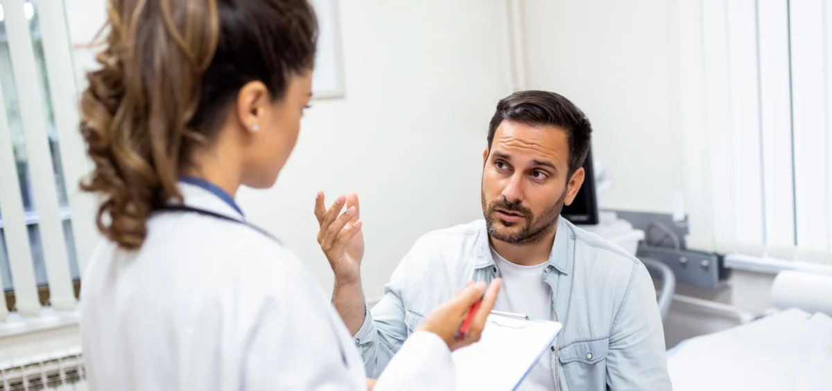 Eine Ärztin steht mit Klemmbrett vor einem jungen Patienten und hört ihm zu.