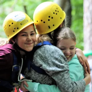 Drei Mädchen in Kletterausrüstung und mit gelben Helmen umarmen sich freudestrahlend.