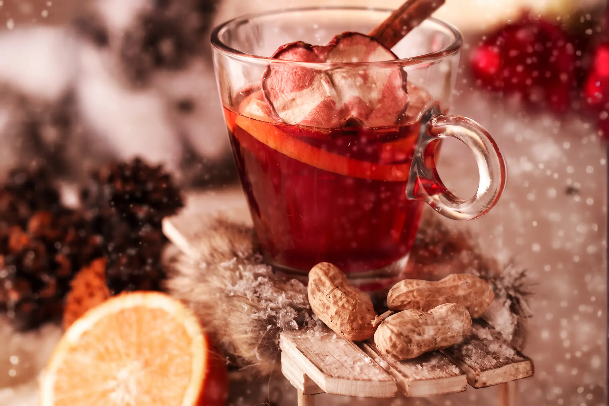 Ein Glas, gefüllt mit Punsch, einer Zimtstange, getrocknetem Apfel und einer Orangenscheibe. Stehend auf einem kleinen Holzschlitten in einem weihnachtlichem Ambiente