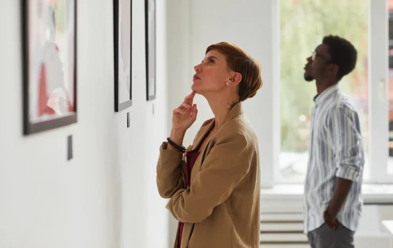 Eine Frau die in einer Ausstellung steht und nachdenklich ein Bild betrachtet. Im Hintergrund steht ein Mann der ebenfalls ein Bild anschaut