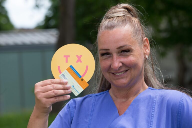 Lisa trägt einen Kasack und hält lächelnd einen Organspendeausweiß und einen TX-Sticker in die Kamera
