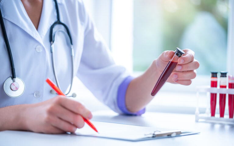 Eine Laborantin/Ärztin hält ein Röhrchen mit einer Blutprobe in der einen Hand, in der anderen einen Stift. Vor ihr liegt ein Klemmbrett. Das Bild symbolisiert Folgendes: Bei Menschen mit chronischer Nierenerkrankung werden wichtige Blutwerte regelmäßig erhoben und kontrolliert.