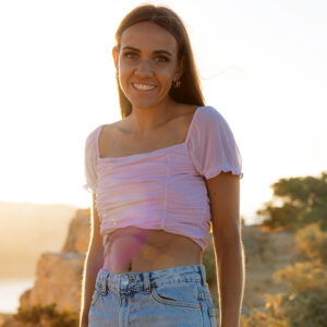 Eine junge Frau bei Sonnenuntergang lächelt in die Kamera. Sie trägt ein bauchfreies Oberteil und hat eine große Narbe am Bauch.