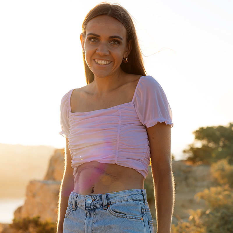 Eine junge Frau bei Sonnenuntergang lächelt in die Kamera. Sie trägt ein bauchfreies Oberteil und hat eine große Narbe am Bauch.