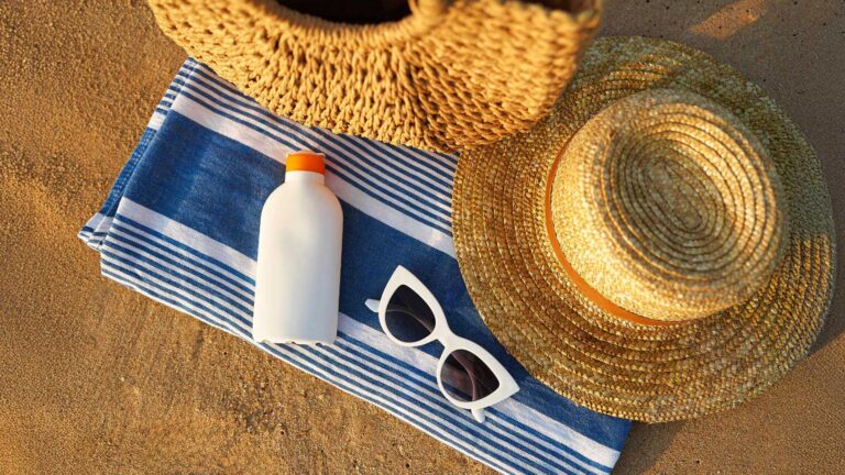 Sonnenschutzmittel, Sonnenbrille und Strohhut auf einem gestreiften Strandtuch im Sand.