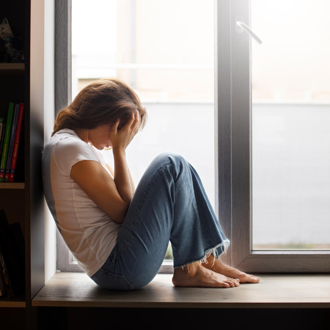 Eine Frau sitzt zusammengekauert auf einer Fensterbank und verbirgt ihr Gesicht in den Händen.